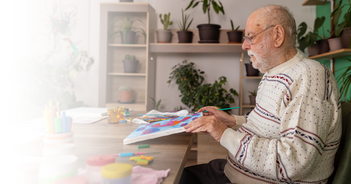 Elderly man with dementia painting at a table in a memory care setting, engaging in a therapeutic, creative activity.